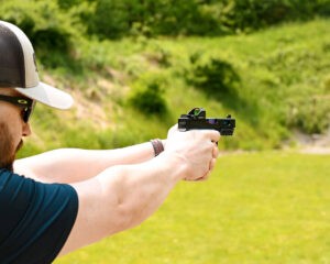Man firing a gun with a red dot sight attached at an outdoor shooting range