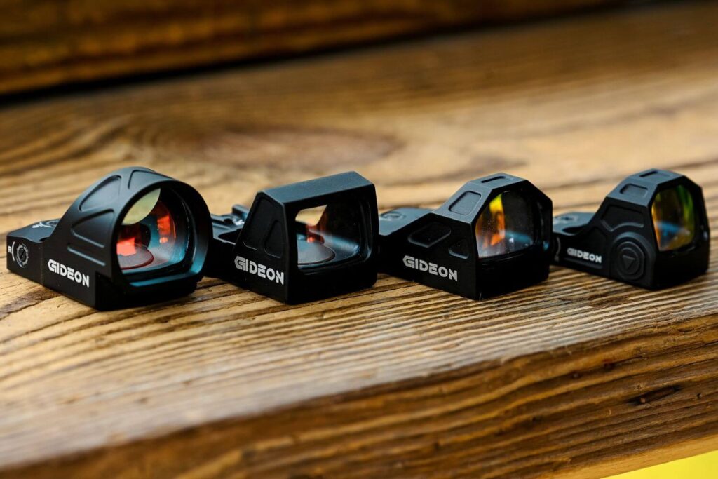 A variety of Gideon Optics red dot sights lined up beside each other on a wooden table