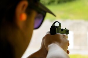 Behind the shoulder view of a man wearing protective earwear aiming a pistol with a red dot sight mounted