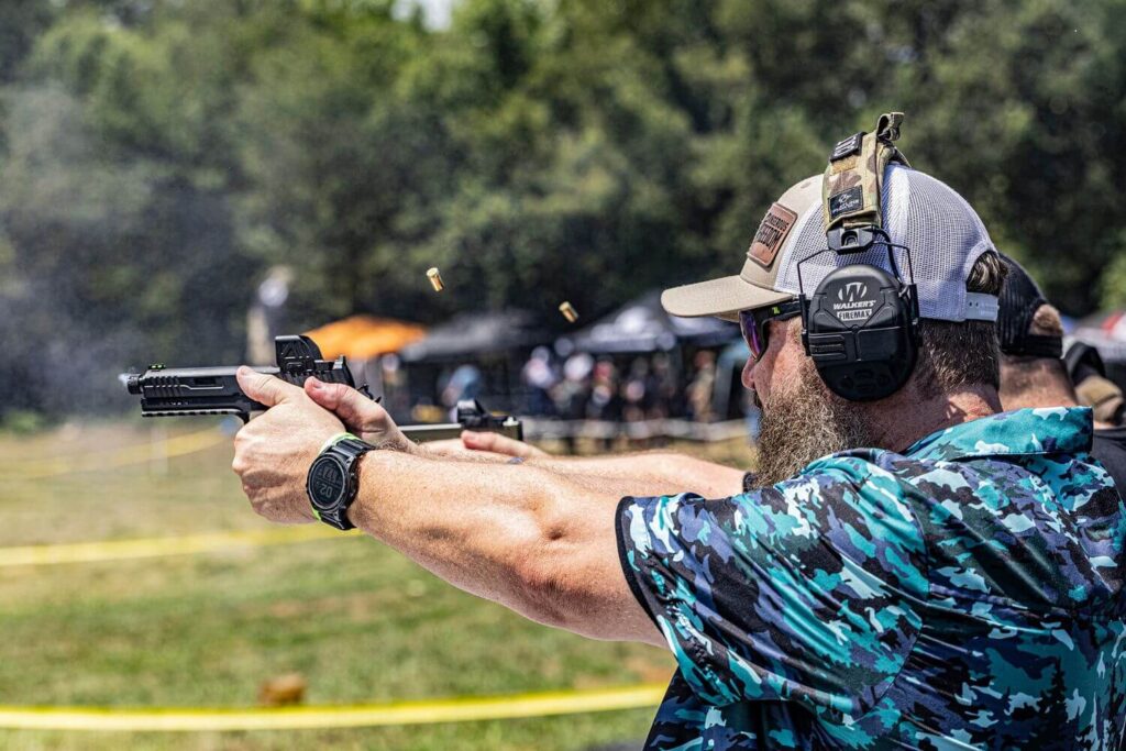 Man wearing blue camouflage shirt and protective eye and ear gear is shooting a pistol with an open emitter red dot mounted