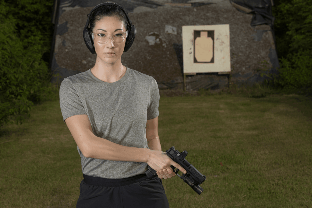 Woman holding a pistol to her side while standing in front of a target at an outdoor shooting range - Gideon Optics Woman holding a pistol to her side while standing in front of a target at an outdoor shooting range