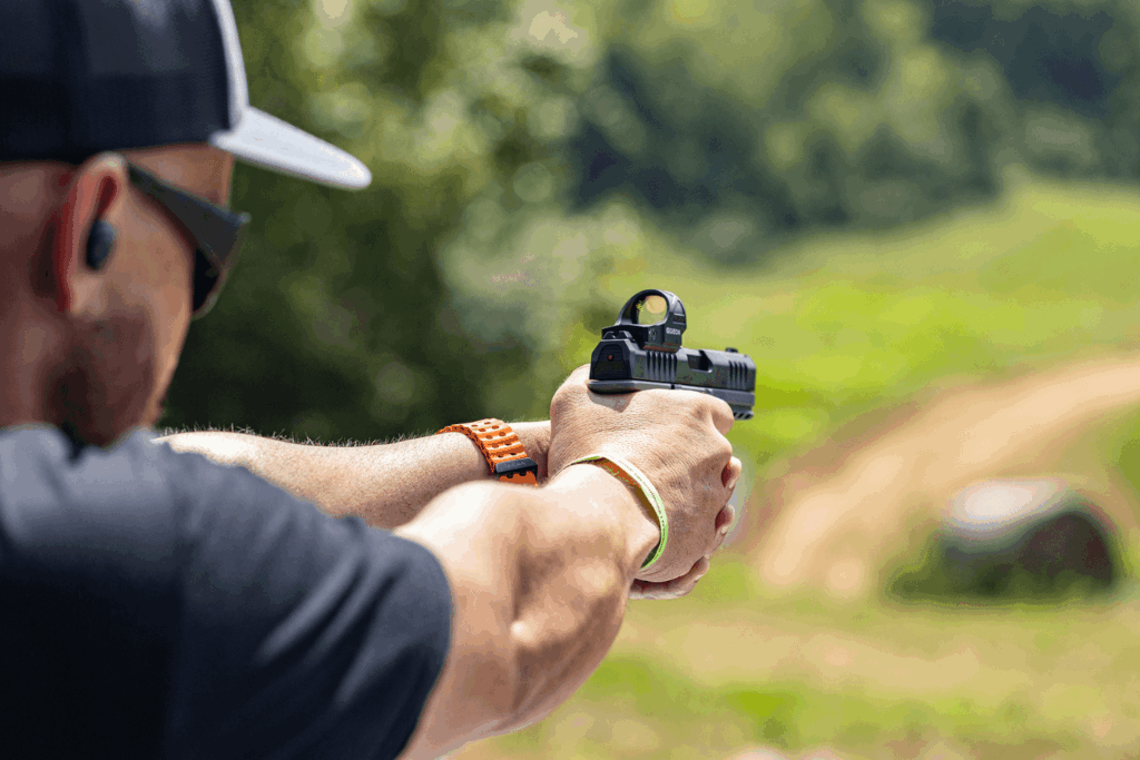 Man wearing a hat glasses and protective earwear firing a pistol at an outdoor shooting range - Gideon Optics Man wearing a hat, glasses, and protective earwear firing a pistol at an outdoor shooting range