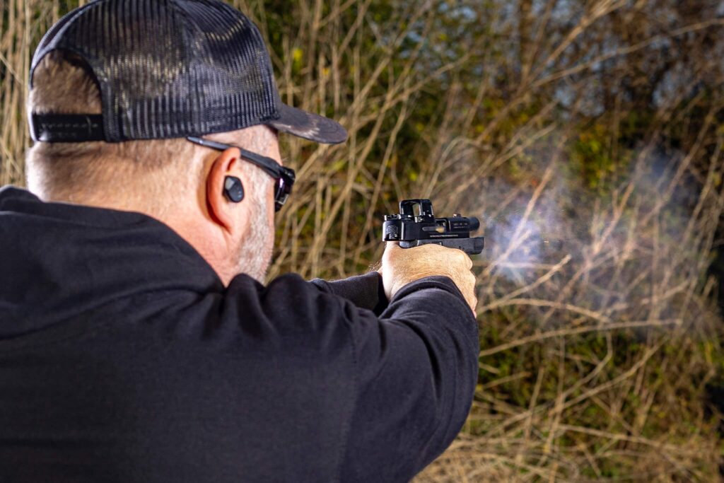 behind the back view of a man in all black at an outdoor shooting range firing a pistol