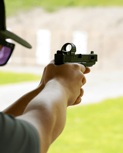 Person wearing sunglasses aims a handgun at a target outdoors, with an extended arm in the foreground.
