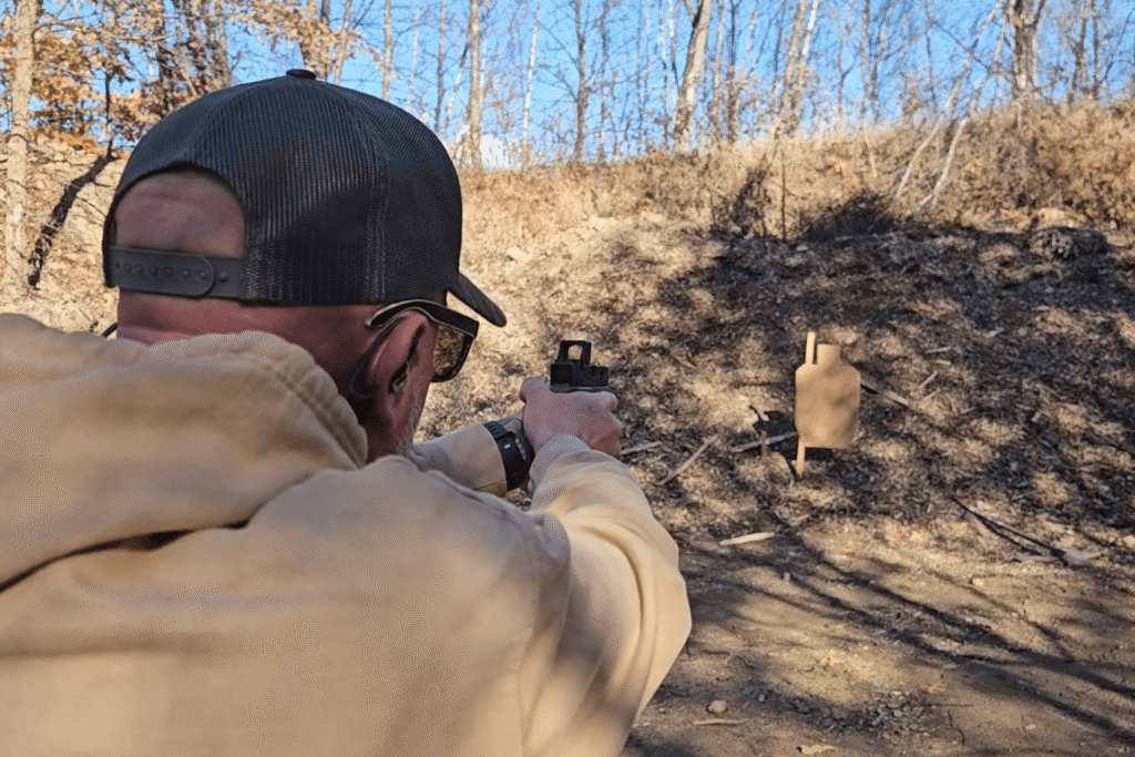Behind the back view of Bob firing his pistol with a red dot mounted at an outdoor shooting range