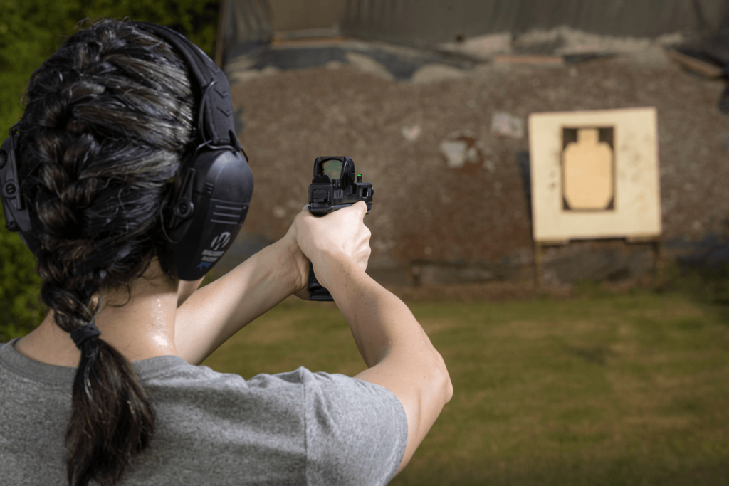 Behind the back view of a woman aiming her firearm with a red dot at a target in the distance