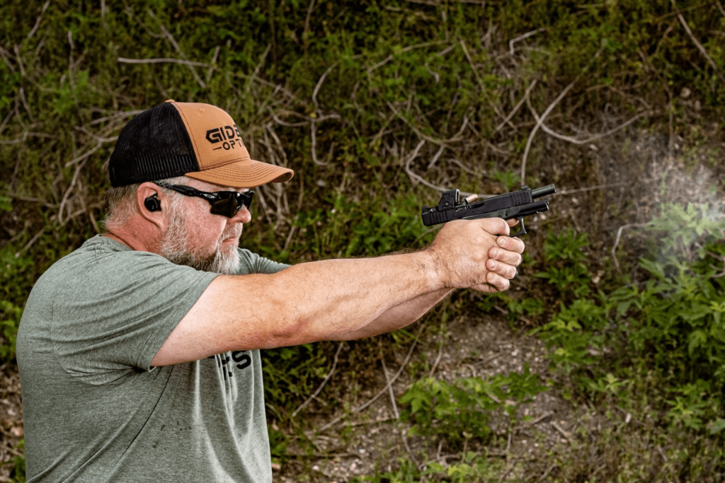 Man wearing a hat and sunglasses firing a pistol with a red dot mounted at an outdoor shooting range