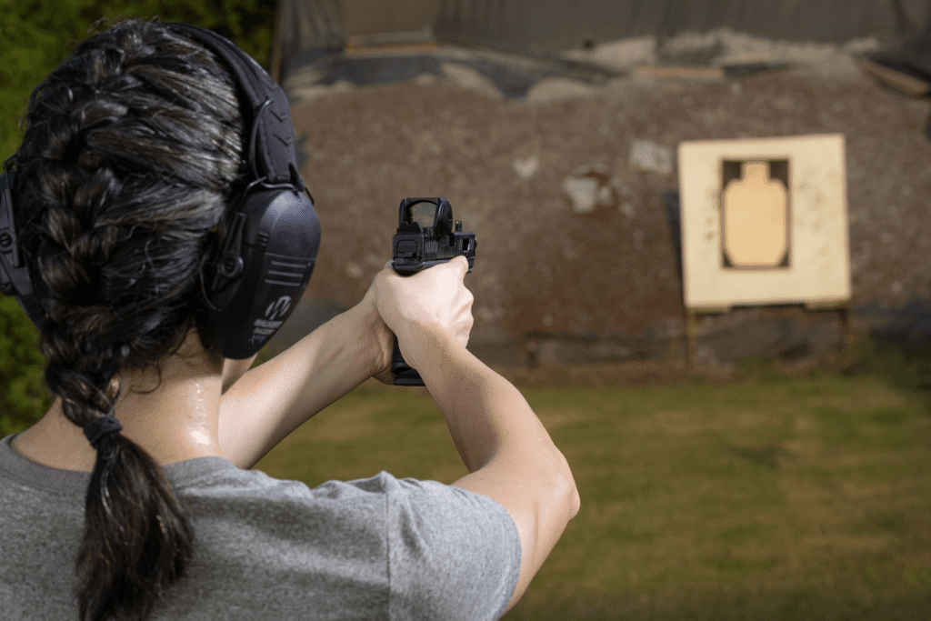 View behind the back of a woman aiming a pistol with a red dot at a target at an outdoor shooting range