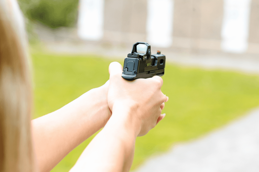 woman shooting pistol - Gideon Optics Close-up of hands holding a black handgun with a red-dot sight, aimed outdoors in a grassy area.