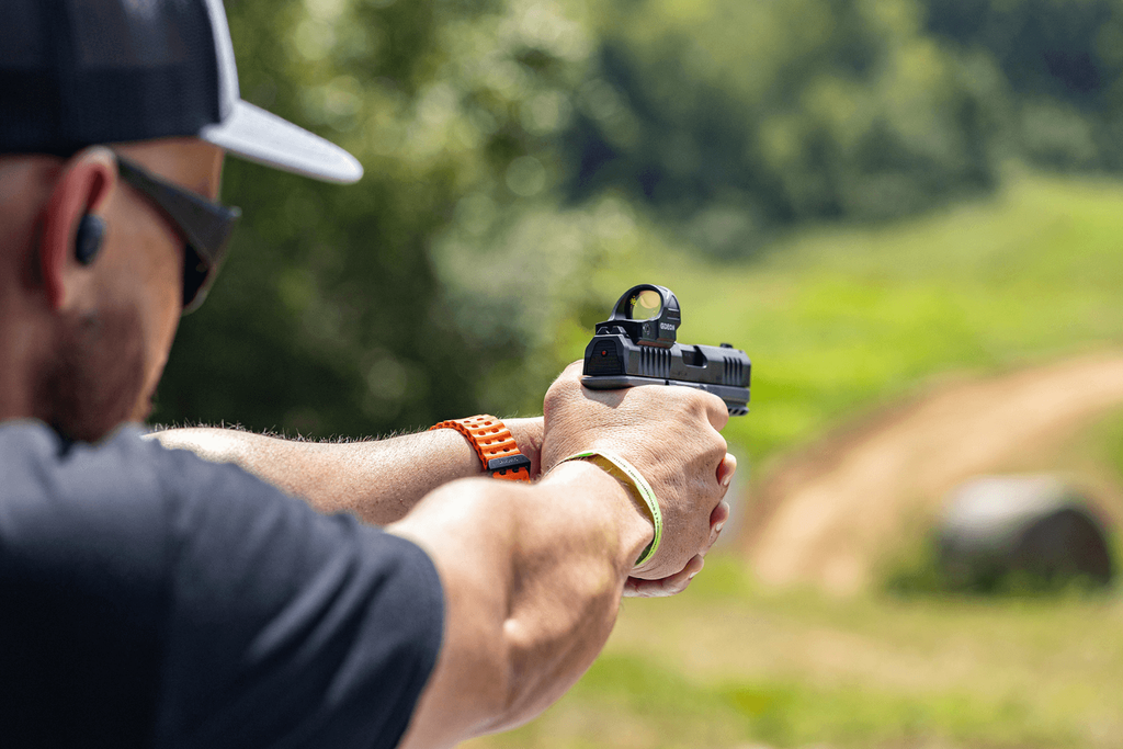 full extension - Gideon Optics Person in a cap aims a pistol with a red-dot sight at an outdoor range or field while wearing a bright orange wristband.